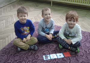 Trzech chłopców siedzi na dywanie. Przed soba mają karty do zabawy