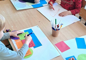 Dzieci siedzą przy stoliku i naklejają kolorowe kartki papieru na biały karton