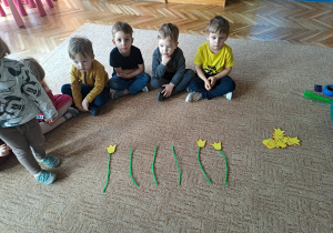 Dzieci siedzą na dywanie obok polozonych papierowych kwiatów