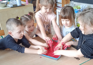 Grupa dzieci wklada ręcę do pojemnika z czerwonym kolorem.