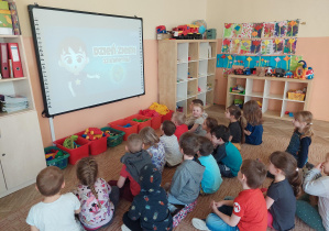 Dzieci siedzą na dywanie i ogladają prezentację.
