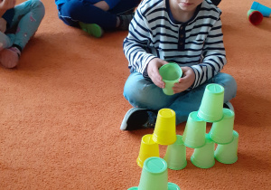 chłopiec siedzi na przeciwko wieży i układa taką samą wieżę z kubków