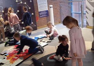 grupa dzieci układa duże puzzle i łapie świecącą gwiazdę przy ścianie z gwiazdozbiorem