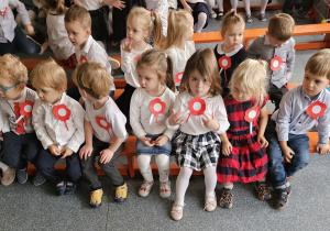 Dzieci siedzą ubrane na galowo z kotylionami biało-czerwonymi