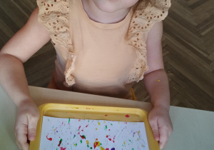 dziewczynka z tacką na której jest kartka z kleksami farby i kulkami
