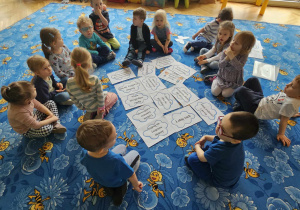 Dzieci siedzą w kole na dywanie a w środku chmurki z napisami