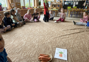 dzieci siedzą na dywanie, na pierwszym planie koszyczek z marchewkami