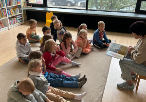 dzieci siedzą na dywanie i słuchają opowiadania czytanego przez panią bibliotekarkę