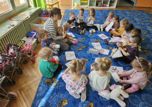 Dzieci siedzą z panią na dywanie i trzymają karteczki ze stworkiem